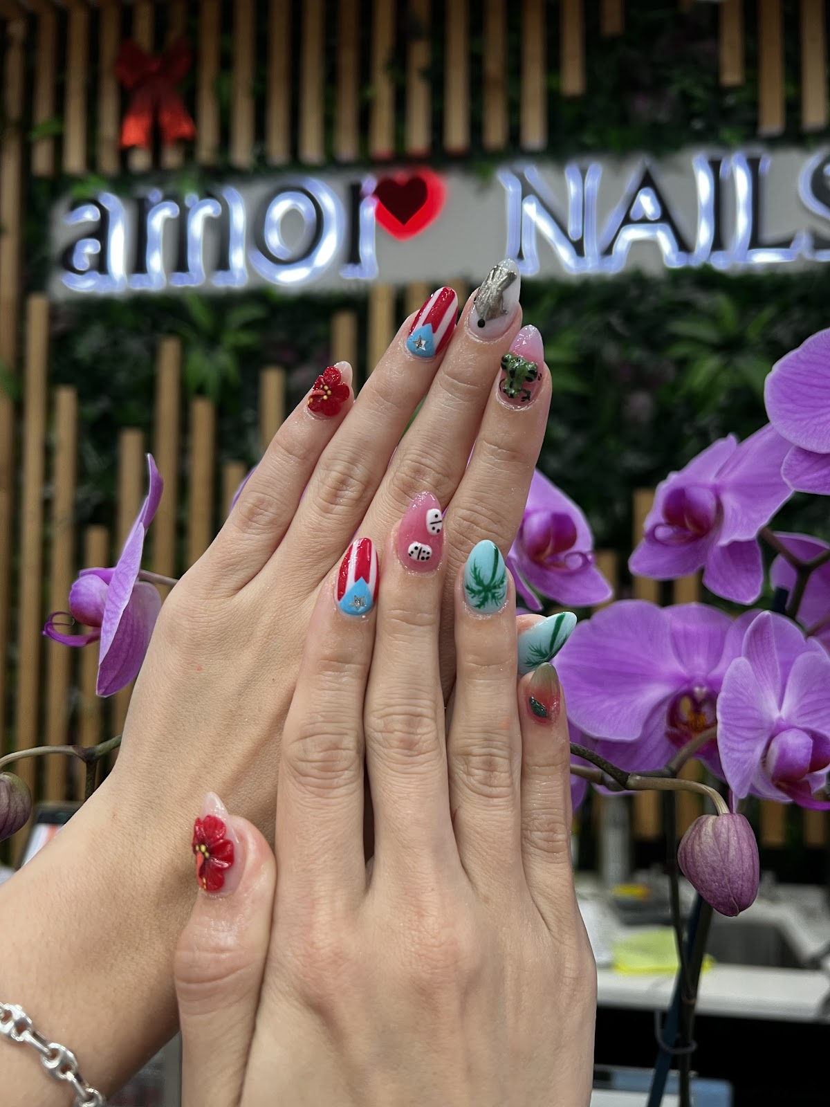 3D floral and rhinestone nail art in front of Amor Nails and Spa's illuminated sign with a heart logo, surrounded by purple orchids